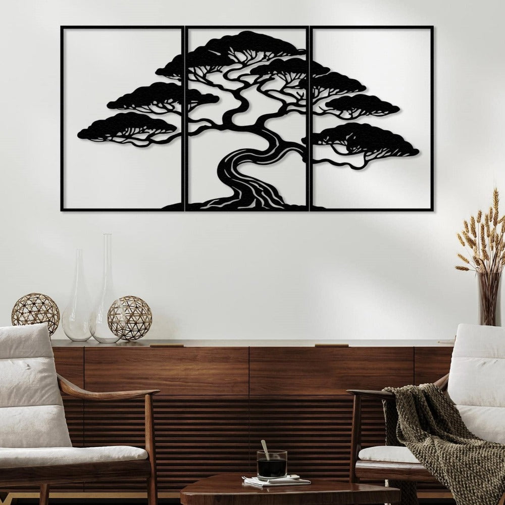 Three-piece black metal wall art featuring a bonsai tree with intricate details, mounted on a white wall above a wooden sideboard. The sideboard is decorated with glass vases and spherical wicker ornaments. The room includes two cushioned wooden chairs and a small coffee table with a glass of coffee and a textured throw blanket.