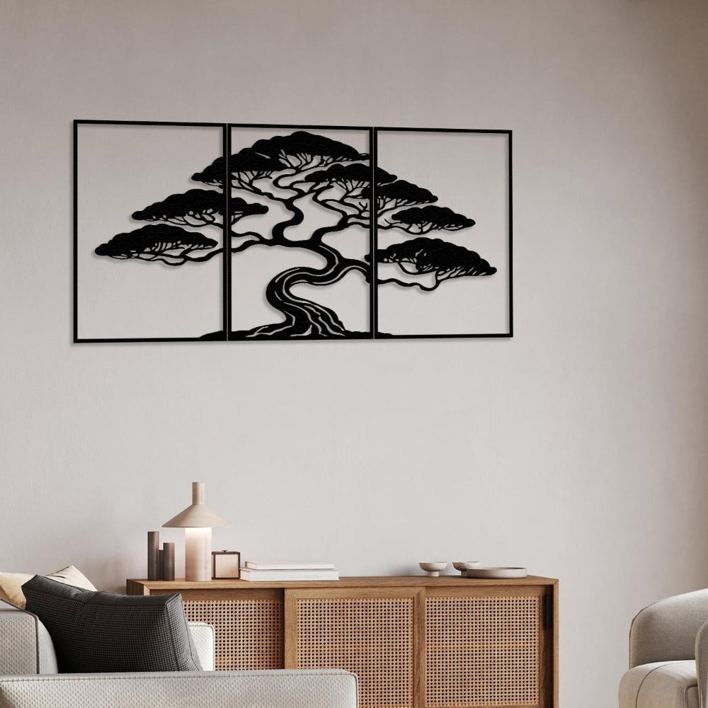 Three-piece black metal wall art featuring a bonsai tree with intricate details, mounted on a light beige wall. Below the artwork is a wooden sideboard with a rattan panel design, decorated with a modern table lamp, books, and small ceramic bowls. The space includes a comfortable sofa with neutral-colored cushions.