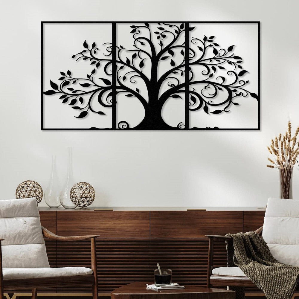 Three-piece black metal wall art featuring a tree with swirling branches and leaves, mounted on a white wall above a wooden sideboard. The sideboard is decorated with glass vases and wicker ornaments, while the room includes two cushioned wooden chairs and a small coffee table with a glass of coffee and a textured throw blanket.