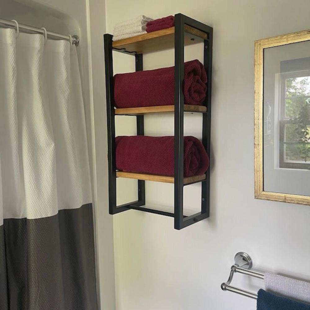 Three-shelf bath towel holder in a modern bathroom setting. The organizer features black metal frames with wooden shelves, neatly holding rolled burgundy towels.