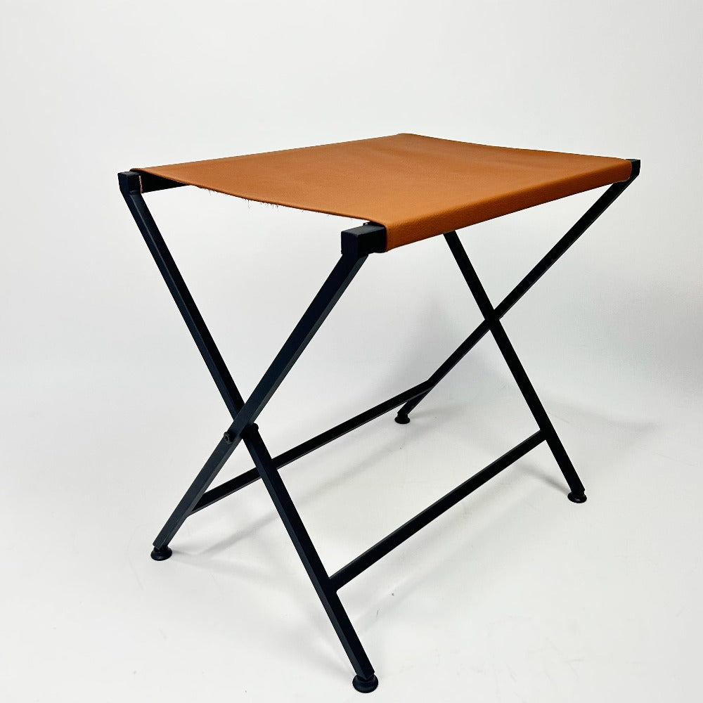 Angled view of a foldable metal suitcase rack, featuring an orange leather top and black frame, highlighting its lightweight and space-saving design.