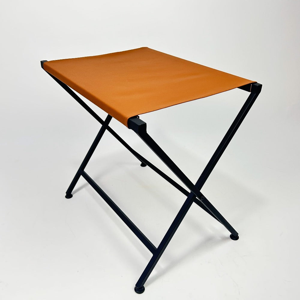 Angled view of a foldable metal suitcase rack, featuring an orange leather top and black frame, highlighting its lightweight and space-saving design