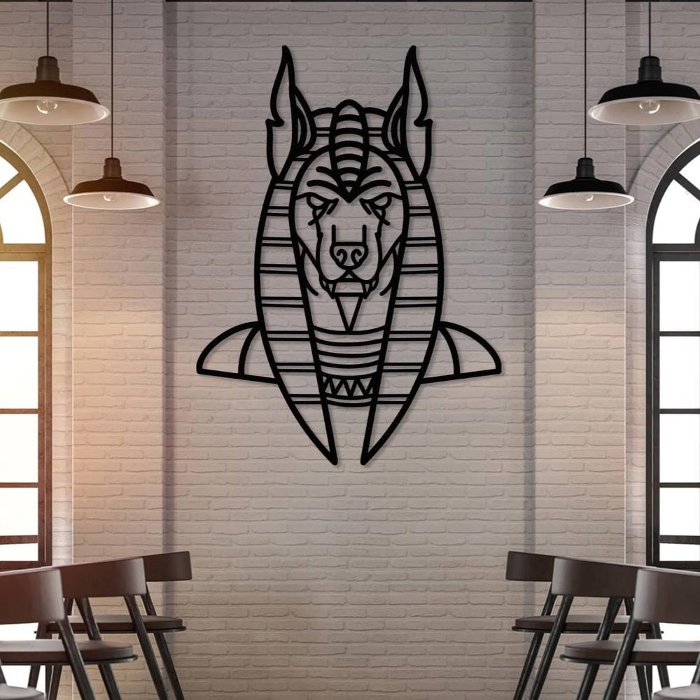 Black metal wall art of Anubis, the ancient Egyptian god, featuring a detailed geometric design, mounted on a light brick wall in a modern bar setting. Industrial-style pendant lights and arched windows frame the space, with wooden chairs arranged below.
