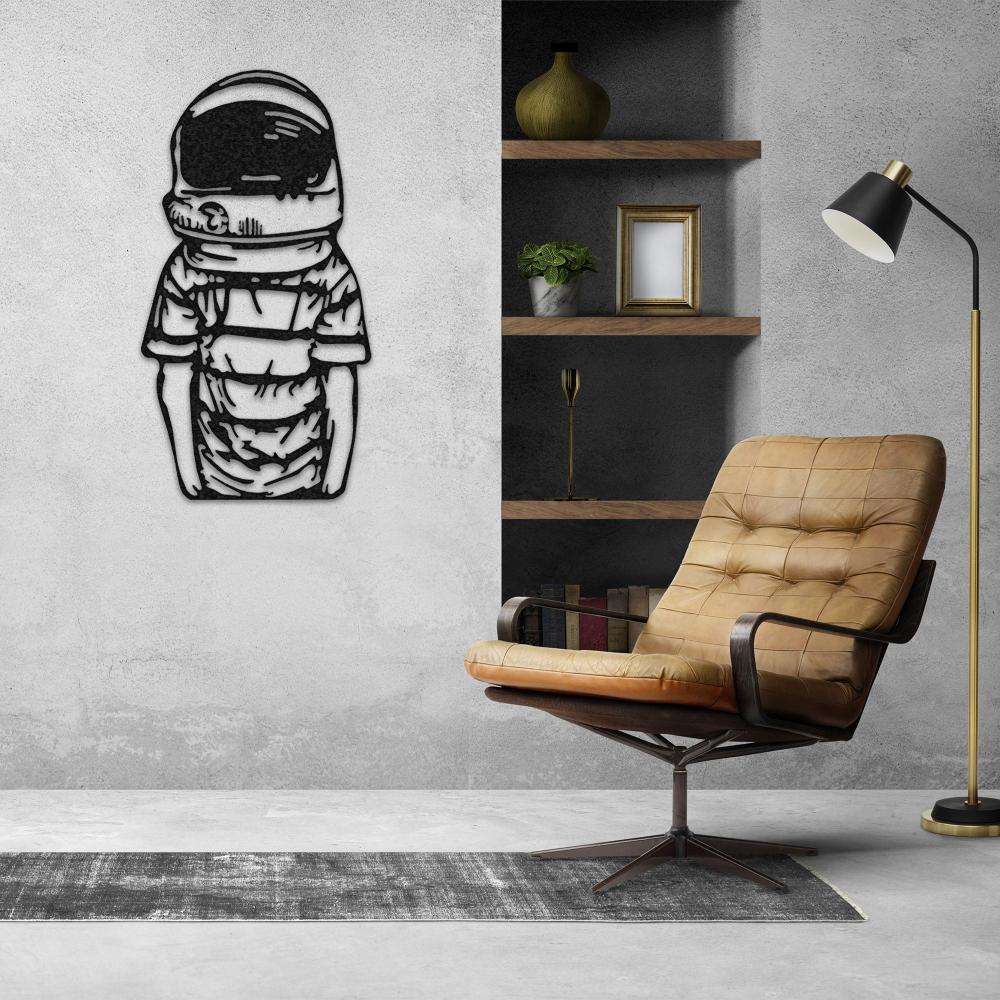 Astronaut-themed black metal wall art featuring a stylized figure in a space helmet and suit, mounted on a modern gray wall next to a cozy leather armchair, wooden shelves, and contemporary decor.