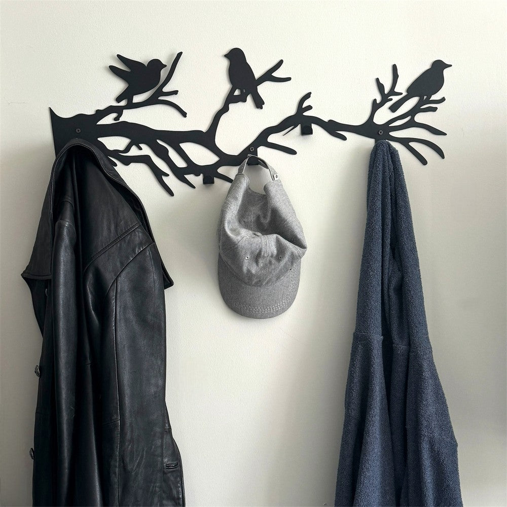 A black metal wall-mounted rack featuring birds on branches, used as a coat hanger. The rack holds a black leather jacket, a grey cap, and a blue towel, combining functionality with a nature-inspired decorative touch.