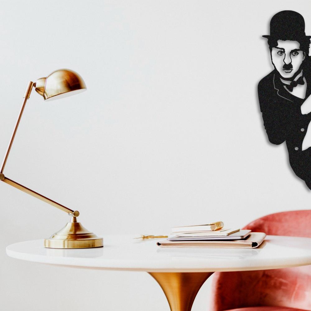 Black metal Charlie Chaplin wall art featuring the iconic figure peeking from behind a corner, mounted on a white wall above a modern workspace with a gold desk lamp, a round white table.