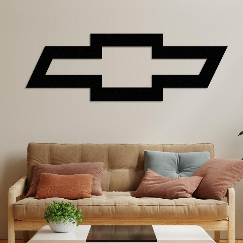 Black Chevrolet-inspired metal car logo wall art displayed on a neutral wall above a beige sofa adorned with rust and blue cushions, accompanied by a small coffee table with a plant centerpiece.