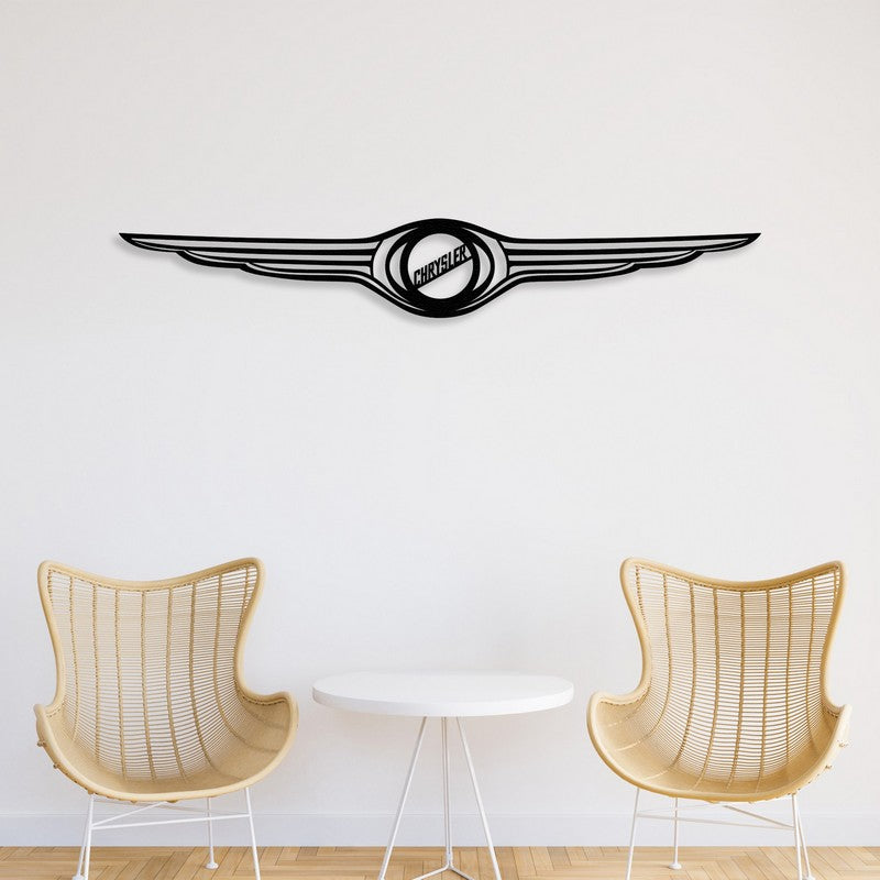 A sleek black Chrysler-inspired metal car logo wall art with a winged design, mounted on a clean white wall. Below, two modern wicker chairs and a minimalist white round table create a stylish and contemporary setting.