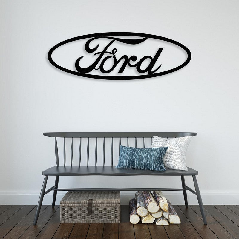 A stylish black Ford-inspired metal car logo wall art mounted on a white wall above a sleek black bench adorned with decorative pillows. The scene includes rustic elements like stacked firewood and a woven basket below, adding a cozy touch.