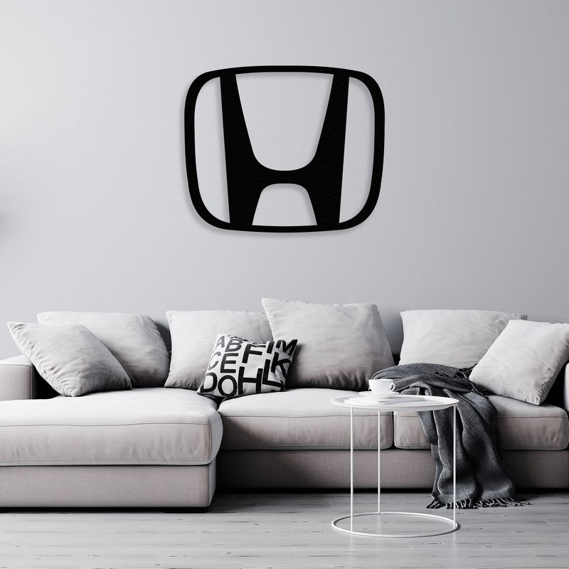 A modern black Honda-inspired metal car logo wall art centerpiece displayed above a light gray sectional sofa with minimalist decor, including monochromatic throw pillows and a small white side table with a cozy throw.