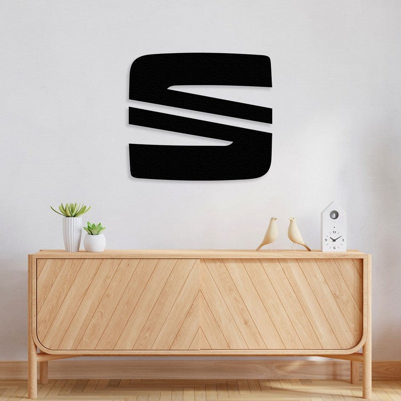 Black metal Seat car logo wall art mounted on a minimalist white wall, placed above a light wooden console with a chevron pattern. The console is decorated with small potted plants, ceramic bird figurines, and a modern white clock, creating a clean and contemporary look.