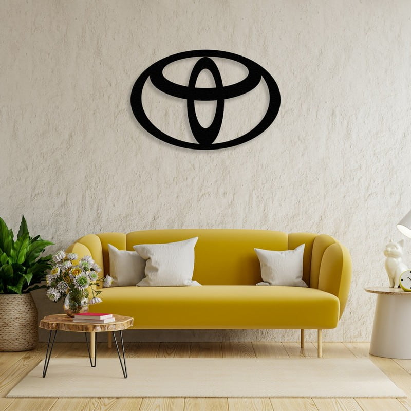 Black Toyota-inspired metal car logo wall art mounted on a textured beige wall above a modern yellow sofa with white cushions, accompanied by a wooden coffee table and indoor plants.