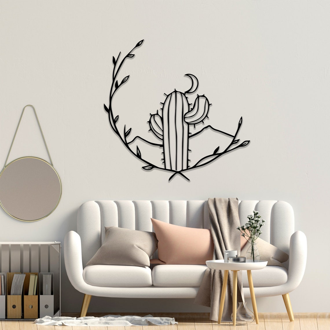 Metal wall art featuring a cactus with a crescent moon and surrounding vines, mounted on a light beige wall above a modern tufted sofa with soft pastel cushions and a throw blanket.
