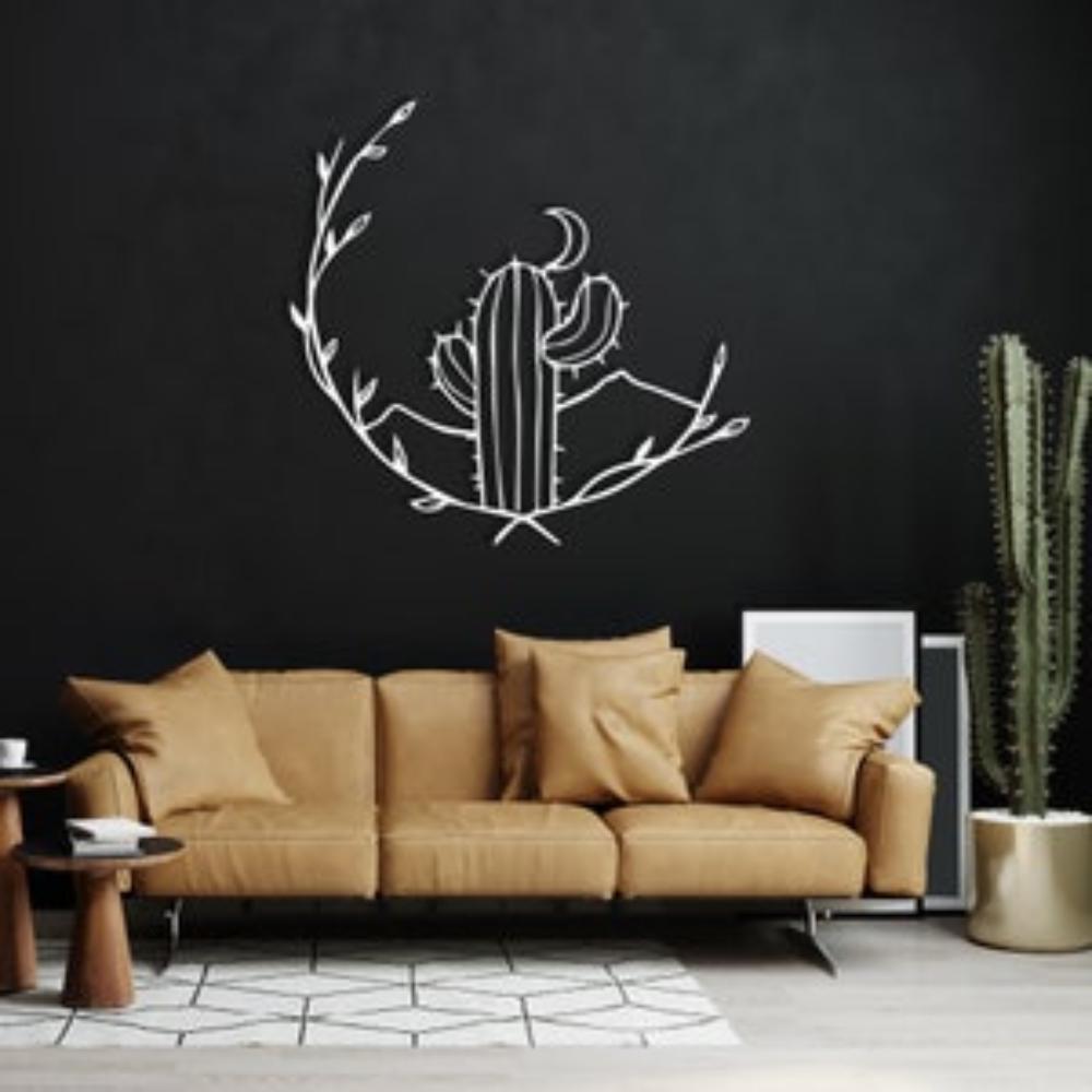 White metal cactus wall art featuring a crescent moon and vines, mounted on a dark wall above a tan leather sofa with beige cushions. A large potted cactus stands nearby, creating a desert-themed living room decor.