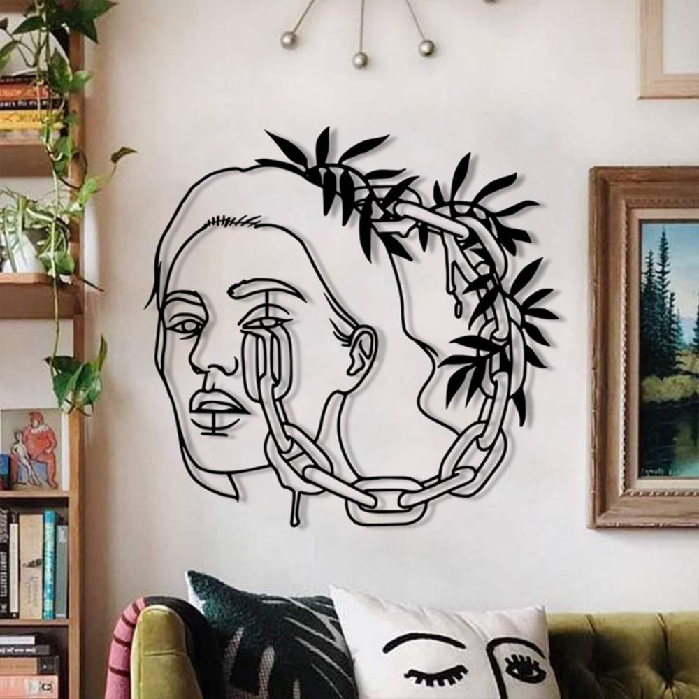 A stylish living room featuring a black metal wall art of a woman's face entwined with chains and leaves, mounted on a light wall with bookshelves and a framed painting.