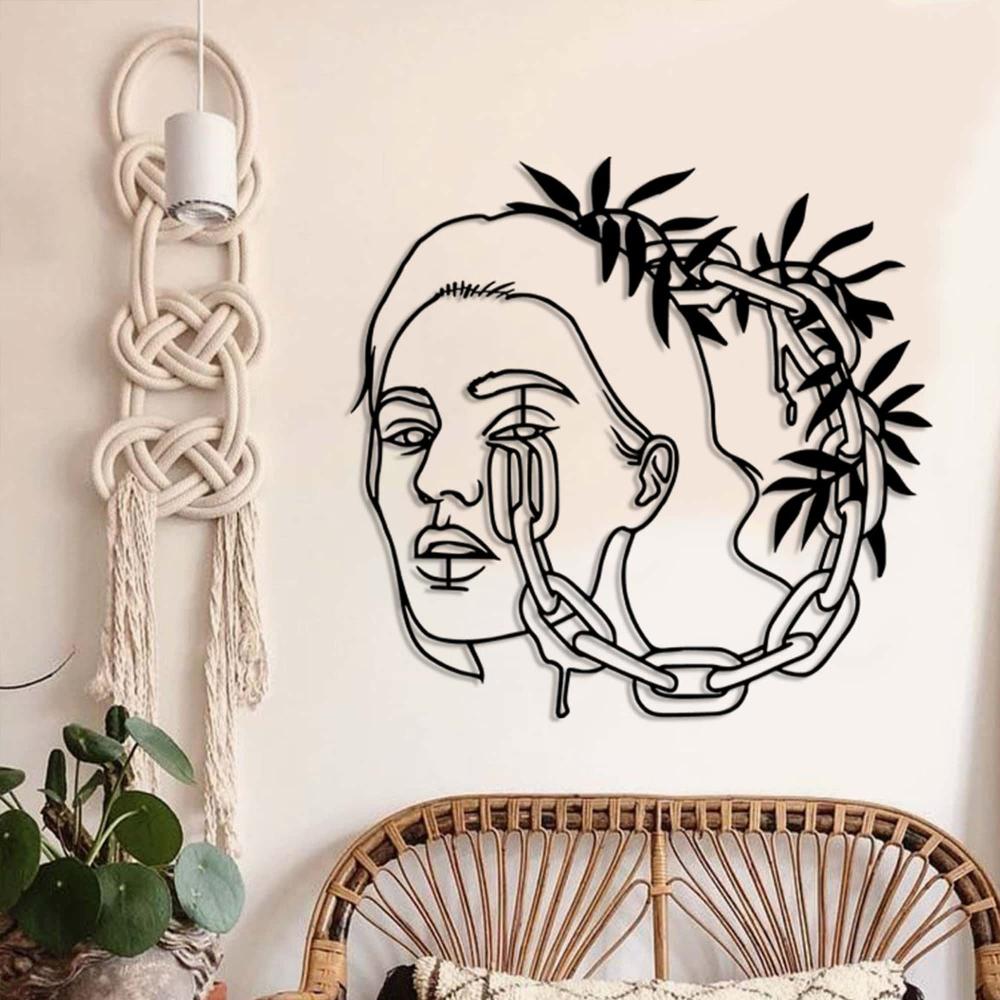 A boho-inspired room with a black metal wall art of a woman's face with chains and leaves, hung next to macrame decor and a wicker chair with a patterned cushion.