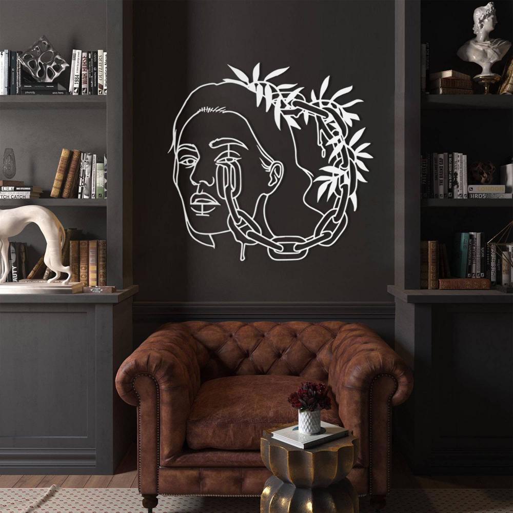 A luxurious library with a dark wall showcasing a white metal wall art of a woman's face surrounded by chains and leaves, placed above a vintage brown leather armchair.