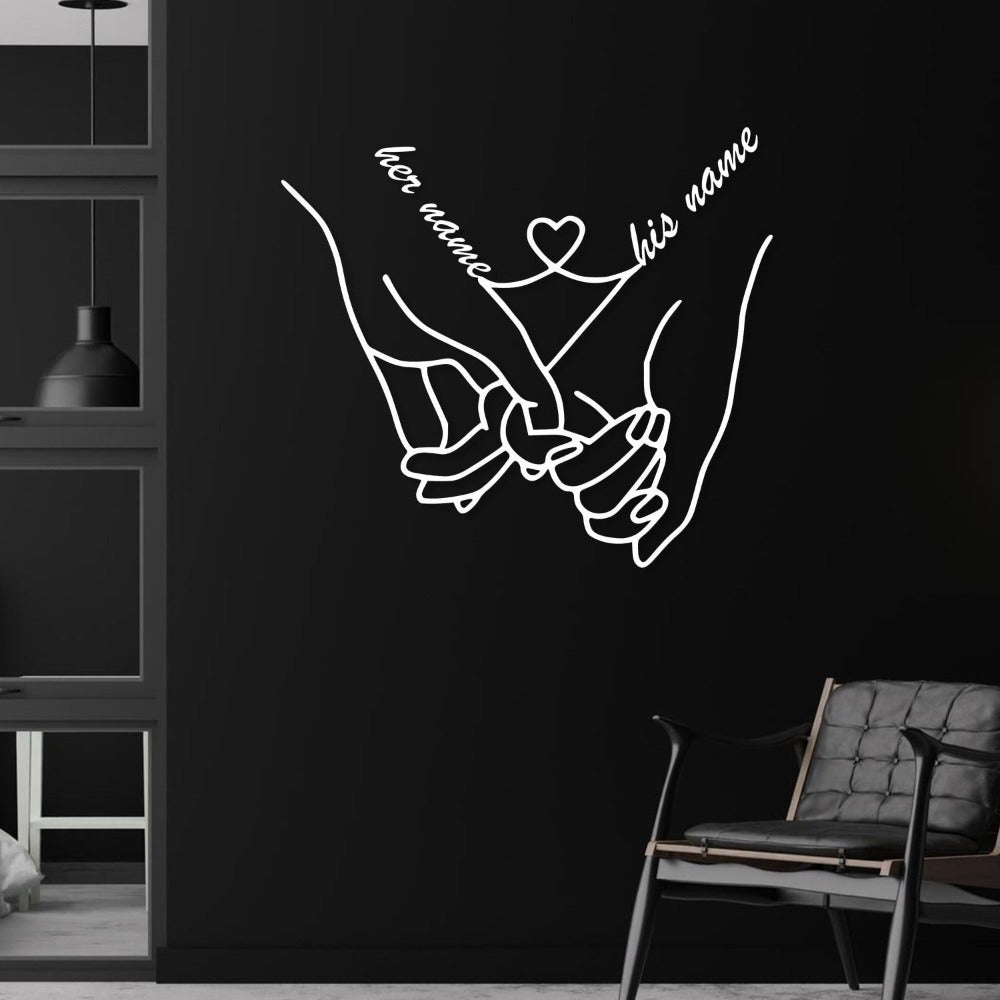 Minimalist metal wall art featuring a pair of hands holding each other, personalized with "her name" and "his name" connected by a heart, elegantly displayed against a dark wall, creating a striking contrast.