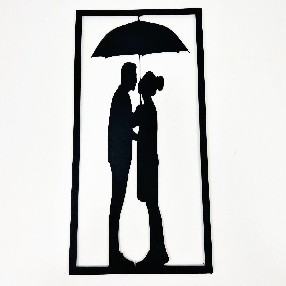 Close-up of black metal wall art featuring a silhouette of a couple standing under an umbrella in a rectangular frame.