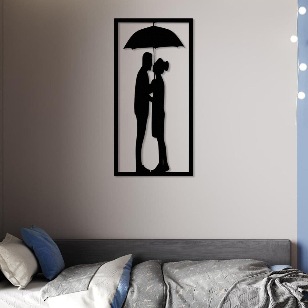 Black metal wall art of a couple standing under an umbrella, mounted above a modern bed with gray bedding and blue and white pillows, softly illuminated by nearby string lights.