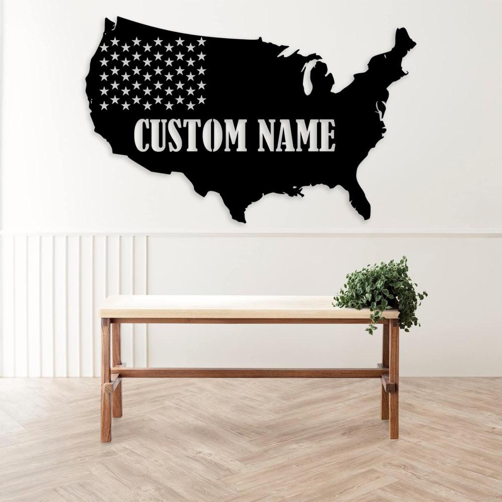 Black metal wall art of the USA map with the text "Custom Name" mounted above a wooden table in a bright space, decorated with a small potted plant.