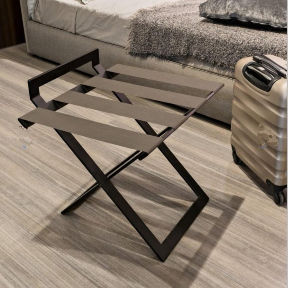 A black luxury luggage rack with wide leather straps placed in a stylish guest room with a modern aesthetic, alongside a beige suitcase on a light wood floor