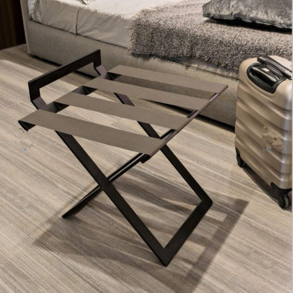 A black luxury luggage rack with wide leather straps placed in a stylish guest room with a modern aesthetic, alongside a beige suitcase on a light wood floor