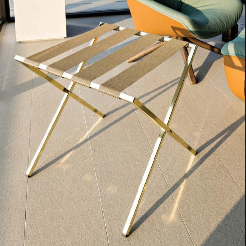 A gold-colored foldable metal luggage rack displayed on a bright patio, featuring elegant brown straps and a practical design