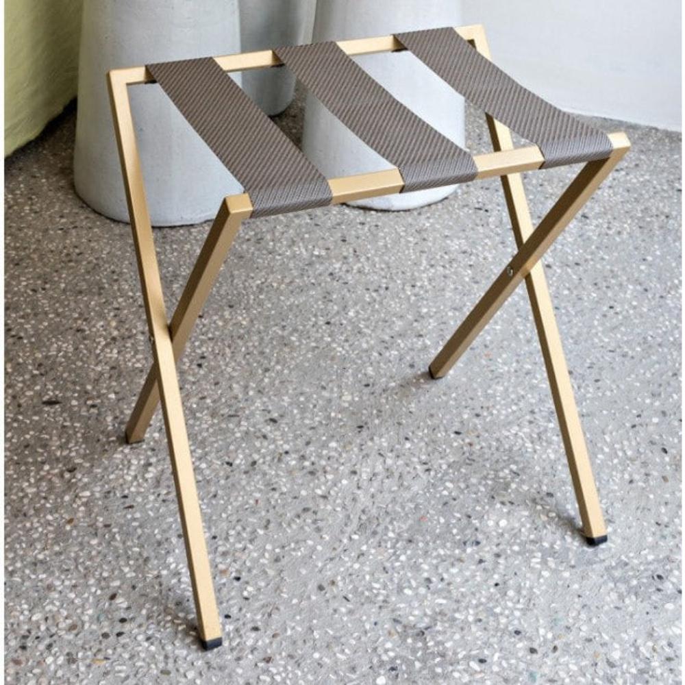 A gold-colored foldable metal luggage rack with brown fabric straps placed on a terrazzo floor, showcasing a sturdy and minimalist design.