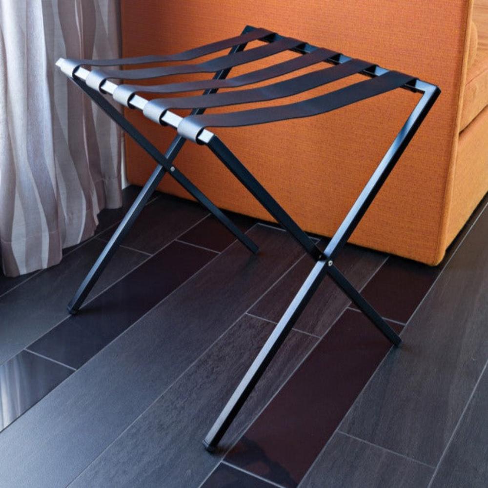 A bronze-tone foldable metal luggage rack with brown straps, paired with a beige suitcase in a softly lit room