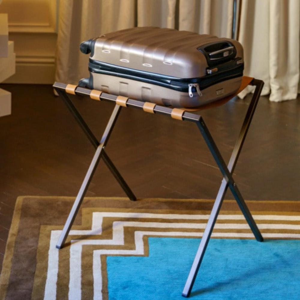 A metal luggage rack with tan leather straps holding a suitcase, positioned on a geometric patterned rug in a stylish interior
