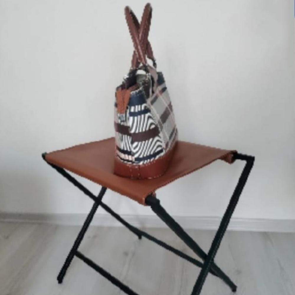 A stylish plaid handbag placed on a foldable metal suitcase rack with a genuine leather surface, standing on a light wooden floor