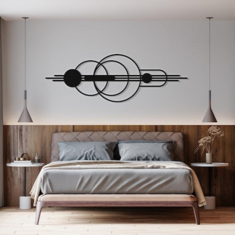 Modern geometric metal wall art featuring intersecting circles and lines, mounted above a minimalist bed with gray bedding and a wooden headboard, flanked by hanging pendant lights and bedside tables in a serene bedroom setting.
