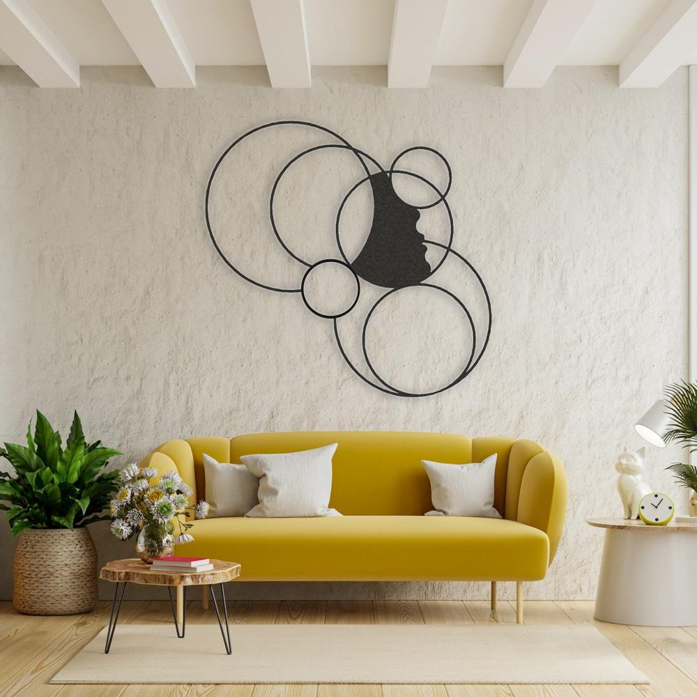 Black metal wall art featuring a girl's silhouette surrounded by overlapping circles, mounted on a textured beige wall above a modern yellow sofa with white cushions, accompanied by a wooden coffee table and indoor plants.