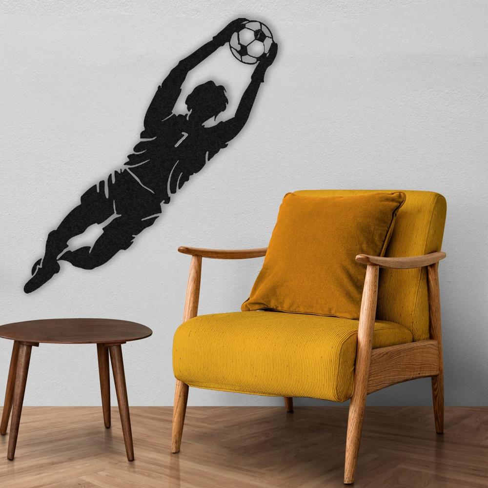 A stylish corner with black metal wall art of a goalkeeper diving to catch a soccer ball, mounted on a white wall above a mustard yellow chair and wooden side table.