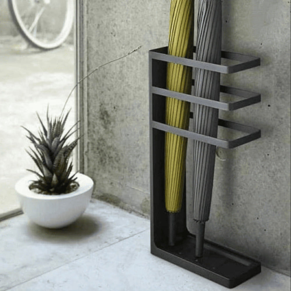 A black metal umbrella stand holding two umbrellas, displayed near a window with a modern concrete backdrop and a decorative plant