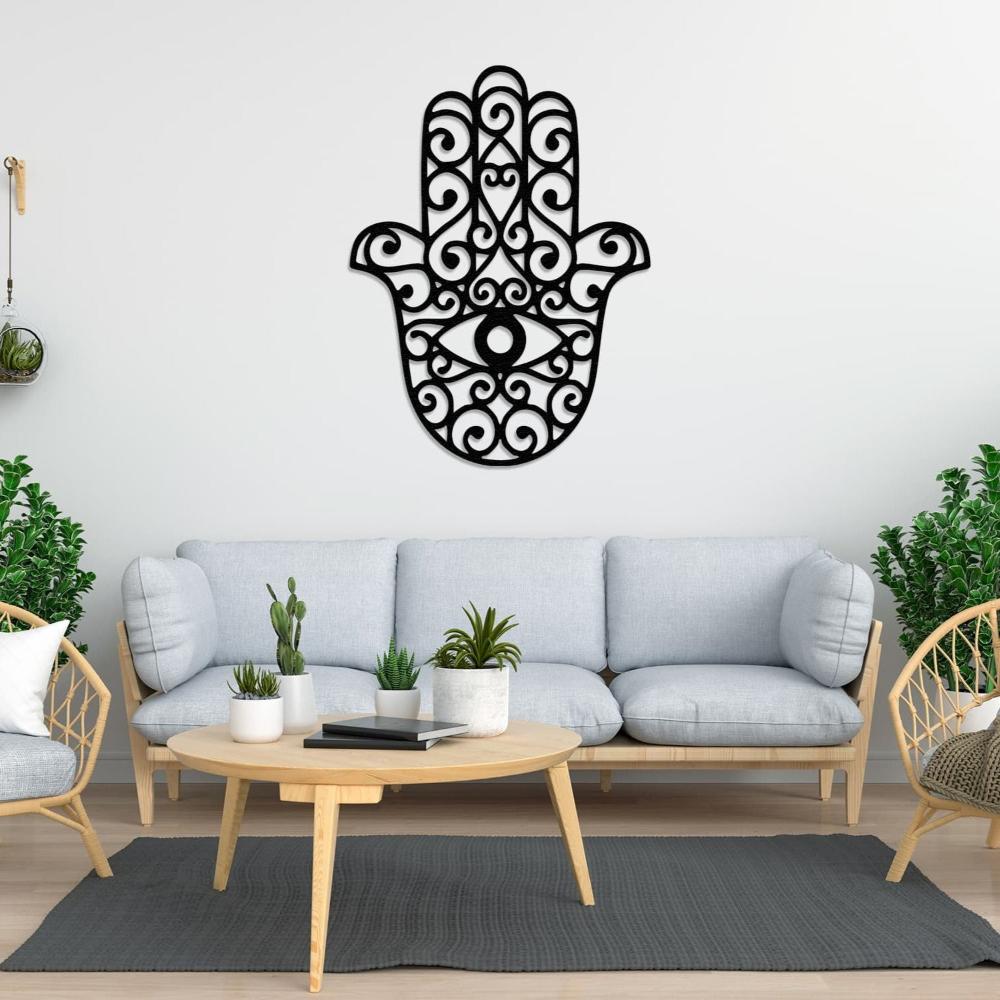 A black hamsa hand metal wall art with intricate swirling patterns and an eye motif, mounted on a light-colored wall above a gray sofa with potted plants in a bright living room.