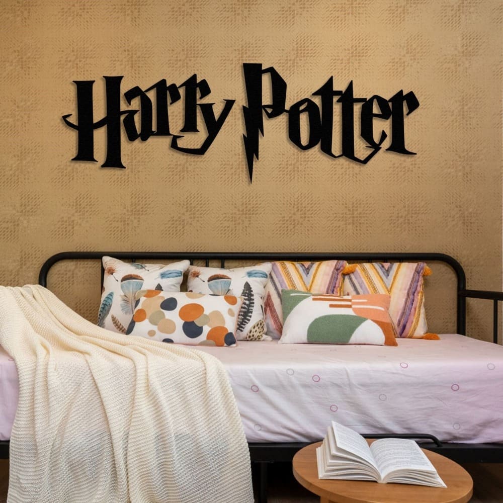 "Harry Potter" black metal wall art with a lightning bolt design, mounted on a textured beige wall above a sofa-style bed adorned with colorful patterned pillows, a white knit blanket, and an open book on a nearby wooden table.