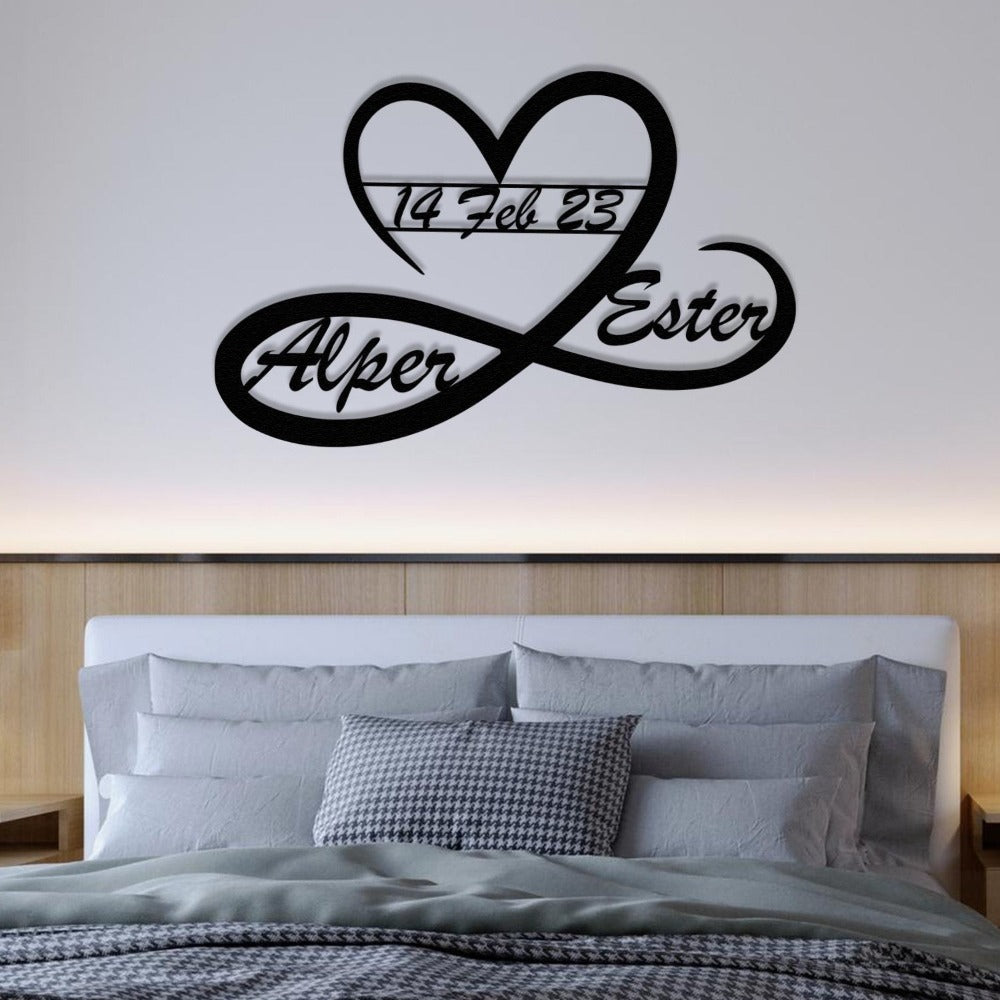 A heart and infinity-shaped black metal wall art featuring the names "Alper" and "Ester" with the date "14 Feb 23" displayed, hung above a modern bed with a grey headboard and bedding.