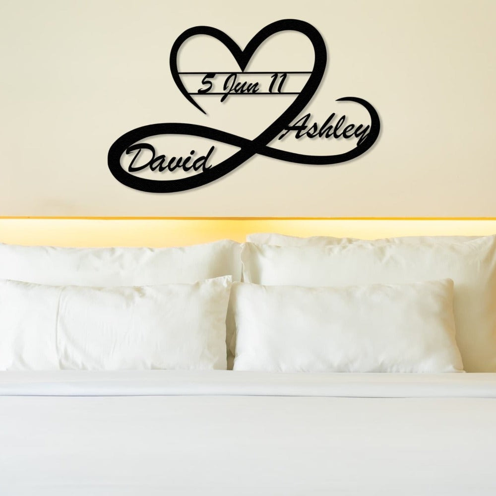 A heart and infinity-shaped black metal wall art featuring the names "David" and "Ashley" with the date "5 Jun 11" displayed above a bed with white pillows and a headboard
