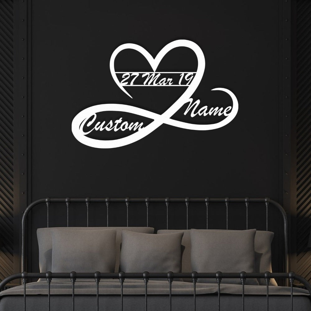 A white heart and infinity-shaped metal wall art with customizable names and date "27 Mar 19," displayed on a dark black bedroom wall above a metal-frame bed with grey bedding.