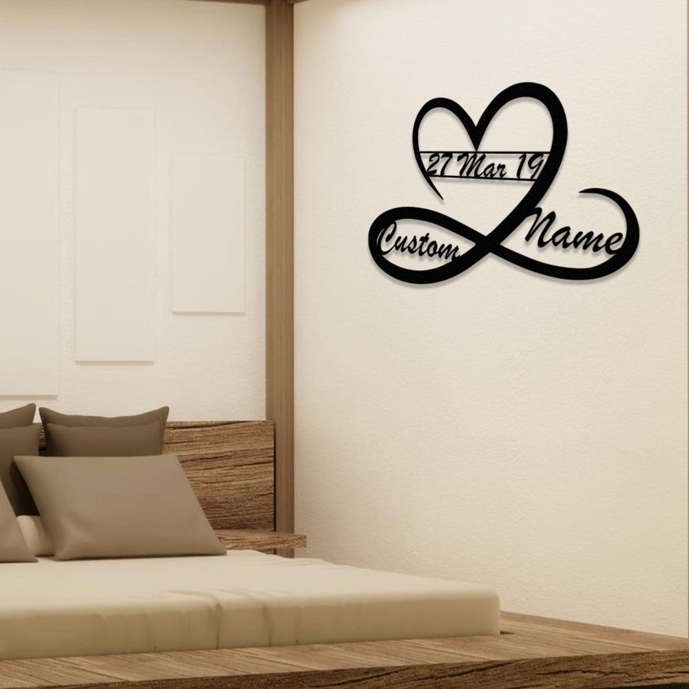 A customized black metal wall art in the shape of a heart and infinity symbol, showing "Custom Name" and the date "27 Mar 19," mounted on a beige wall in a bedroom.