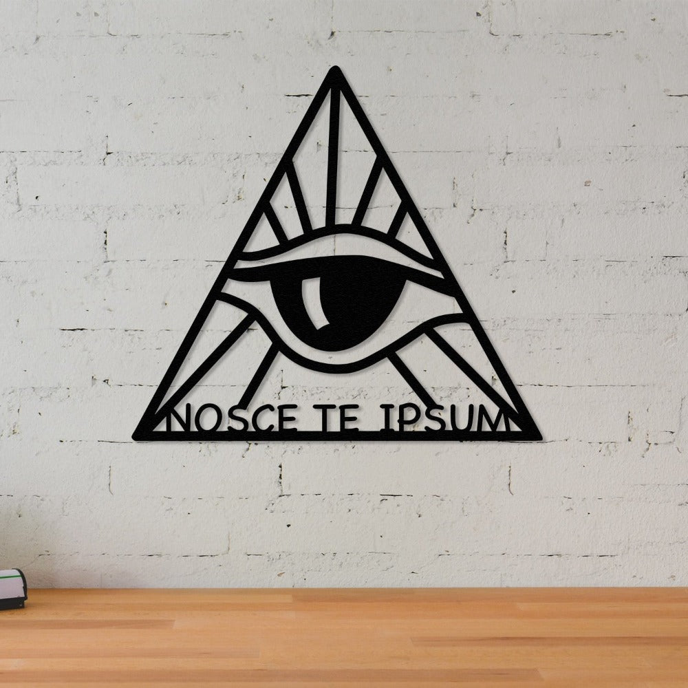 Close-up view of the "Know Thyself" black metal wall art featuring an eye enclosed within a triangle. The phrase "Nosce Te Ipsum" is displayed at the bottom. Mounted on a textured white brick wall above a minimalist wooden tabletop.