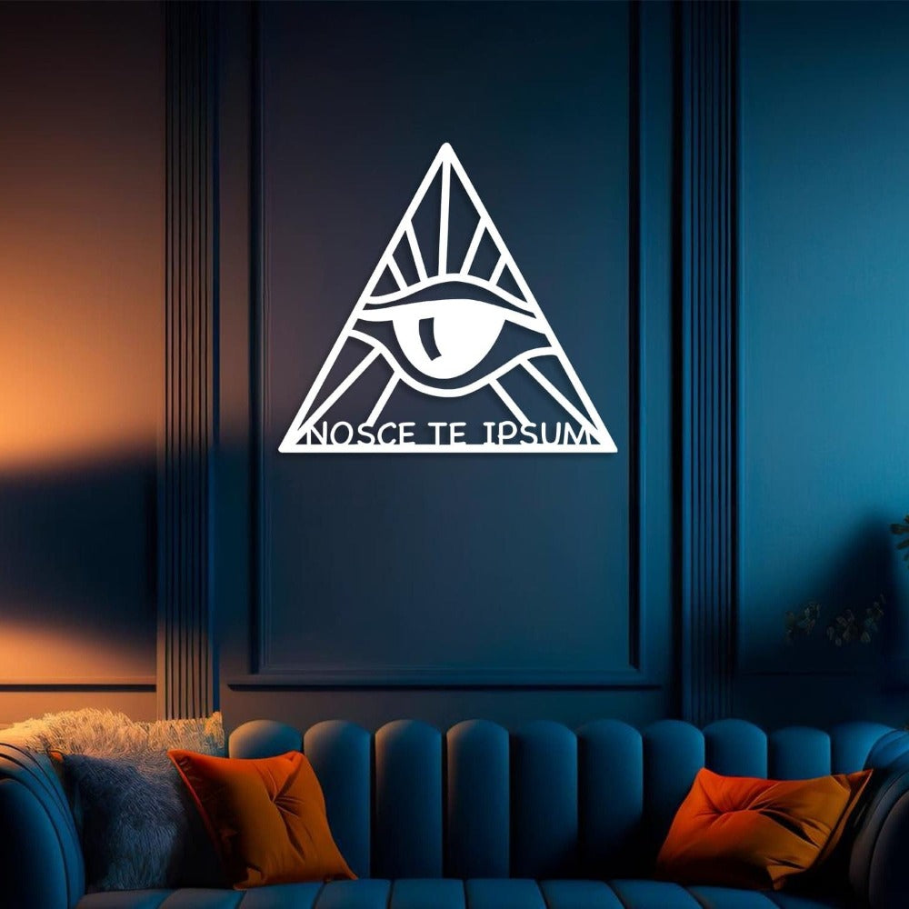 Illuminated living room with a dark blue wall featuring the white "Know Thyself" metal wall art. The design includes an eye within a triangle, emitting an elegant glow, with the phrase "Nosce Te Ipsum" at the base. Positioned above a tufted blue sofa adorned with orange and gray pillows, complementing the ambient lighting.
