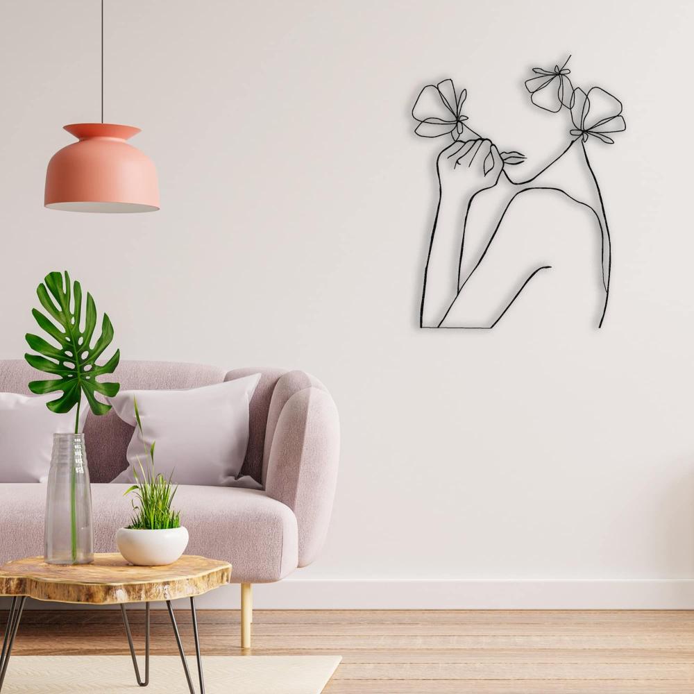 An abstract metal wall art piece of a female figure with flowers, displayed in a contemporary living room with a pastel pink couch and decorative elements.