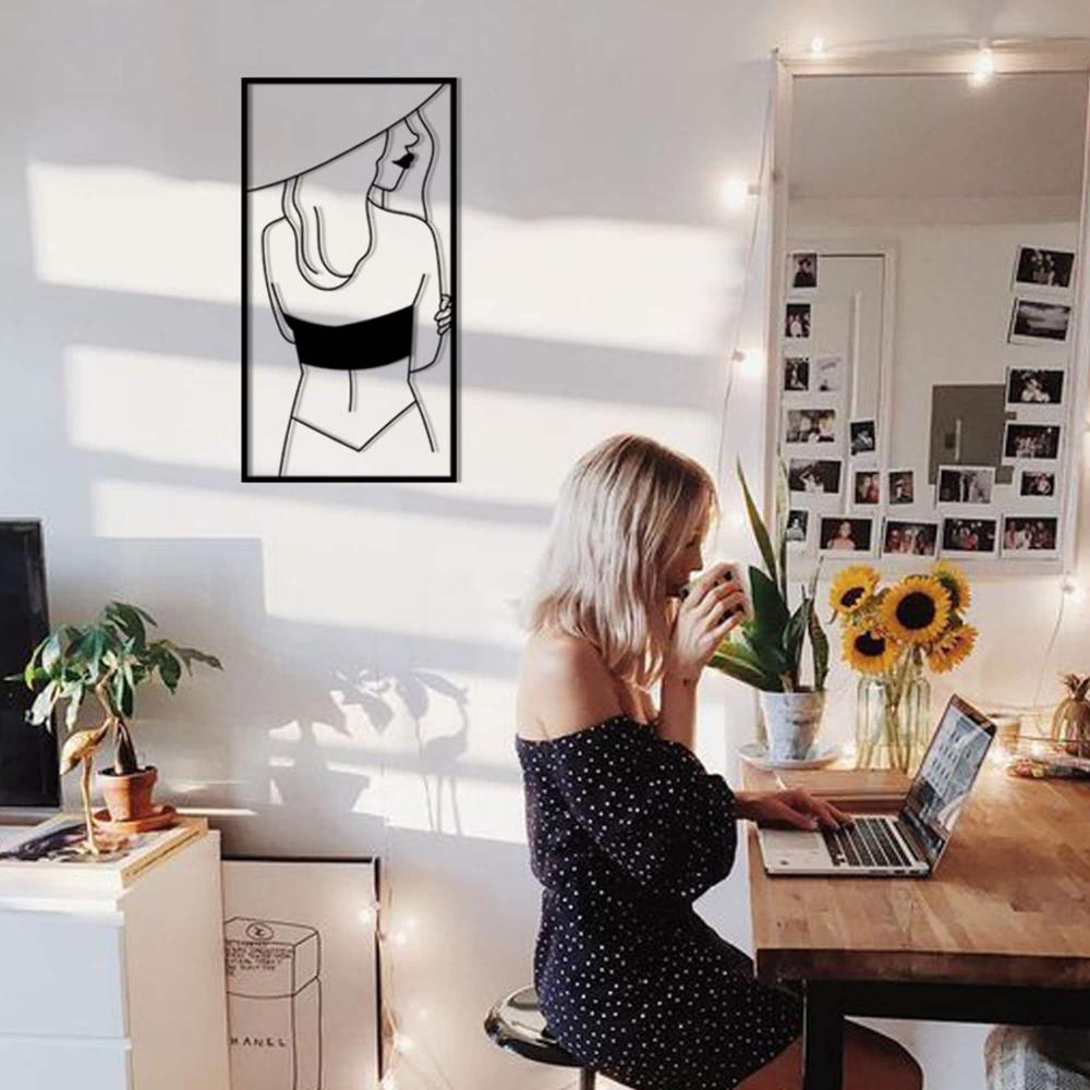 A chic workspace with a woman enjoying coffee and working on her laptop, with a metal wall art of a woman wearing a hat mounted on the wall.