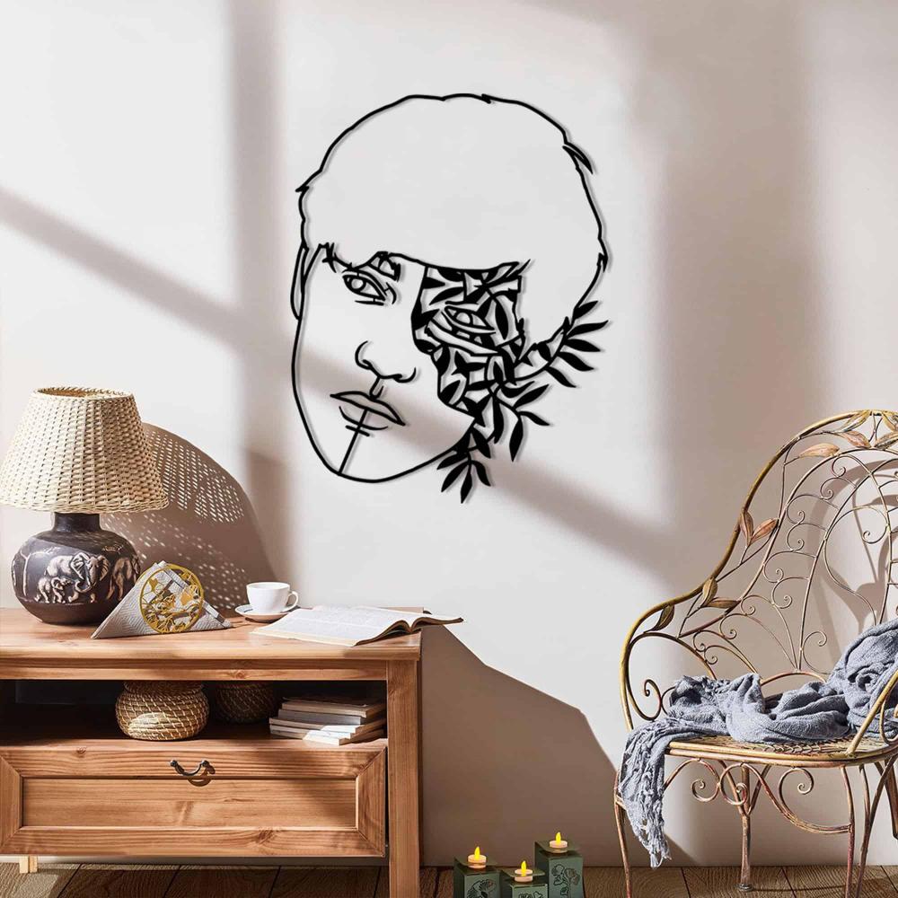 A black metal wall art piece featuring a man's face and floral patterns, mounted on a white wall with sunlight casting shadows, situated above a wooden sideboard with decor items like a wicker chair and candles.