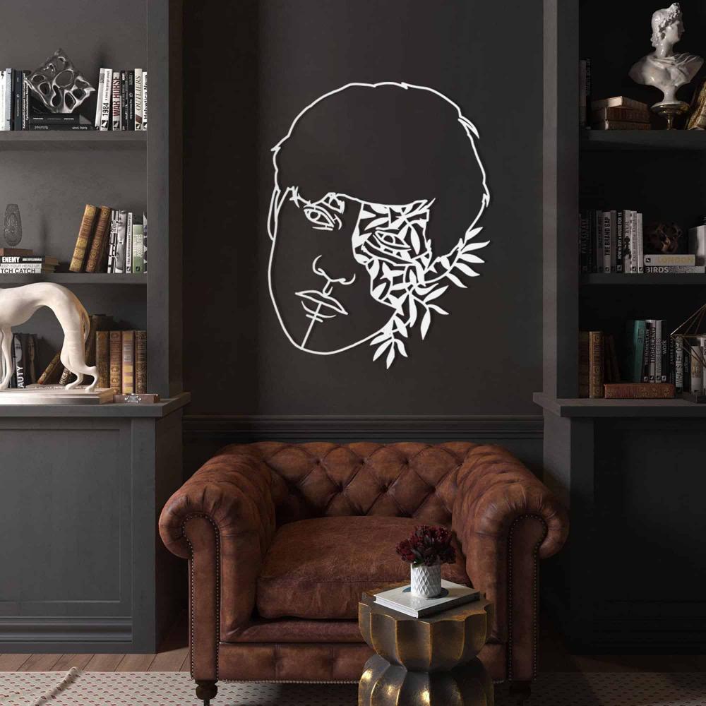 A white metal wall decor of a man's face with floral and leaf elements, mounted on a dark wall in a classic library room with a brown leather armchair and decorative bookshelf.