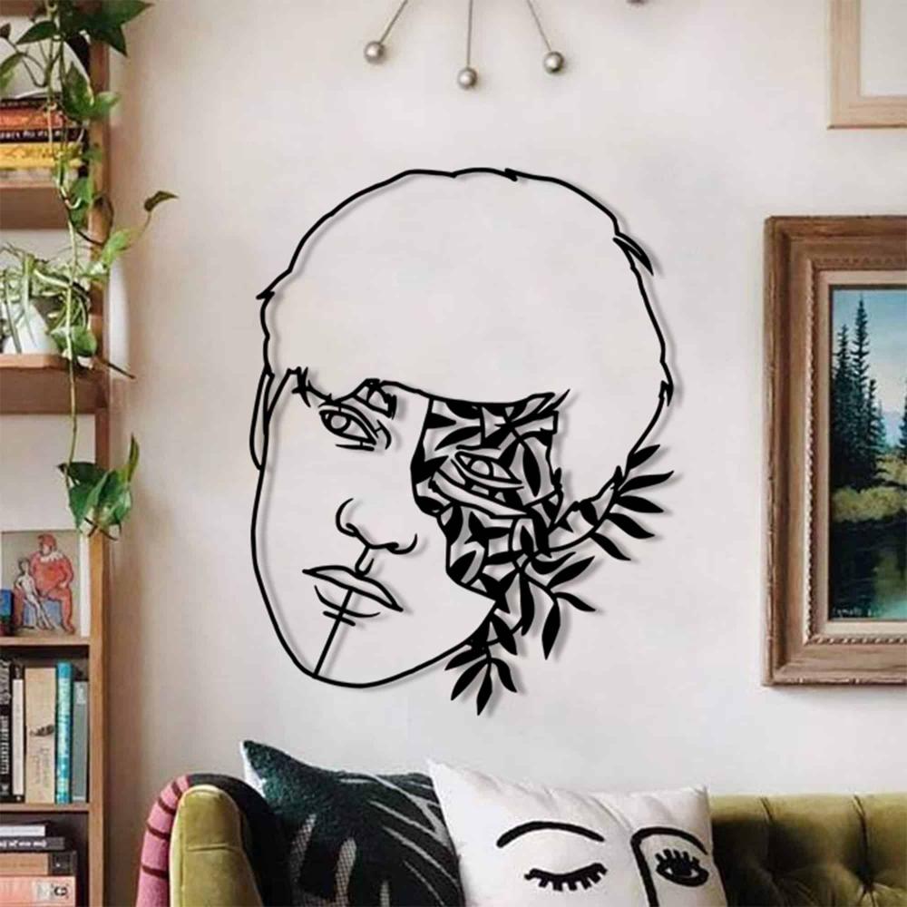 A geometric metal wall art piece featuring a man's face partially overlaid with floral and leaf patterns, mounted on a white wall in a modern living room with wooden furniture and greenery.