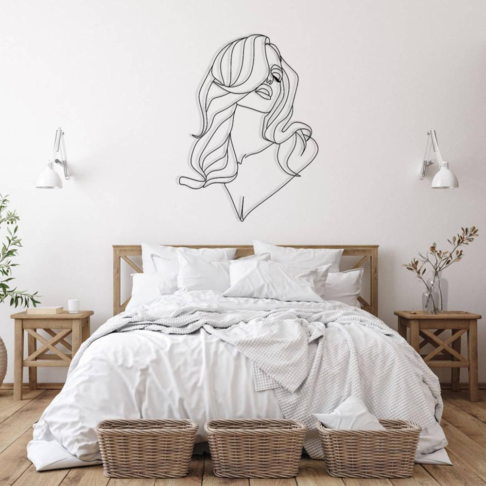 Minimalist line art metal wall decor of a woman with long, flowing hair installed on a white wall above a cozy bed. The bedroom setup includes a wooden bed frame with white bedding, wicker baskets at the foot of the bed, wooden side tables with vases and plants, and white wall-mounted reading lamps.